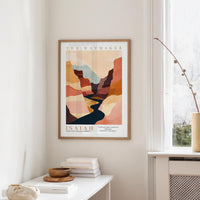 a framed poster of a desert landscape with a river, hanging on a wall in a room with a white table and a window.