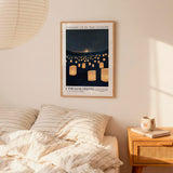 a bedroom scene with a framed poster of a night sky with numerous lit lanterns hanging from the ceiling, a bed with striped bedding, and a wooden nightstand with a coffee mug and a book.