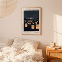 a bedroom scene with a framed poster of a night sky with numerous lit lanterns hanging from the ceiling, a bed with striped bedding, and a wooden nightstand with a coffee mug and a book.