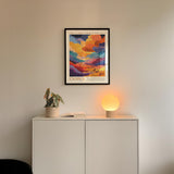 a white cabinet with a plant and a lamp on top, and a framed poster of a colorful landscape hanging on the wall above it.