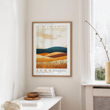 a framed poster of a landscape with a desert scene, hanging on a wall in a room with a window and a small table with books and a bowl on it.