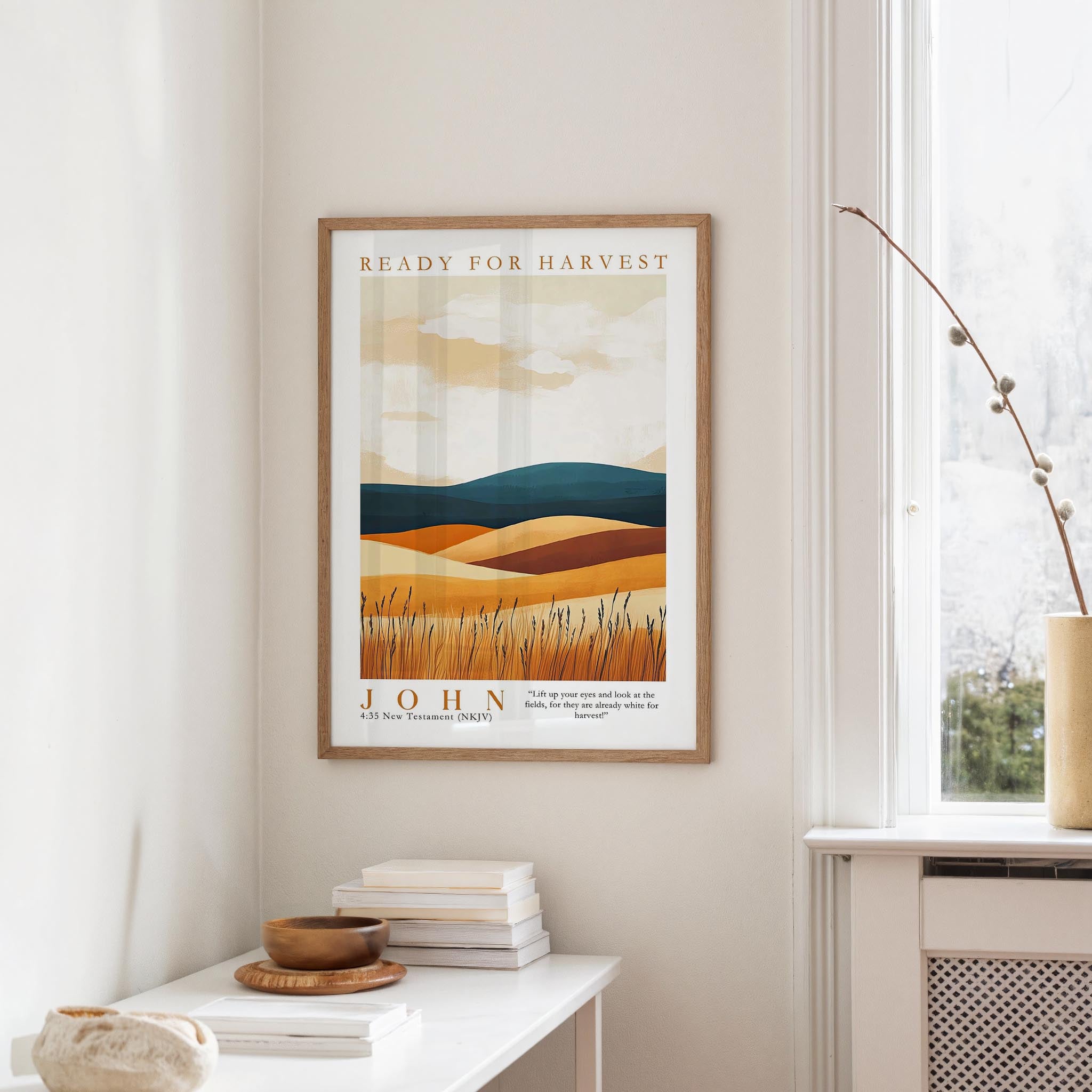 a framed poster of a landscape with a desert scene, hanging on a wall in a room with a window and a small table with books and a bowl on it.