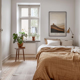 a cozy bedroom with a large bed, a wooden nightstand, and a framed poster on the wall. The room has a warm, inviting atmosphere with a wooden floor and a window overlooking a hillside.