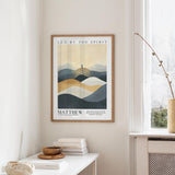 a framed poster of a mountain landscape with a quote from the Bible, "MATTHEW, 'The son of man is the devil' NKJV". The poster is hanging on a wall above a white table with a stack of books and a small vase.