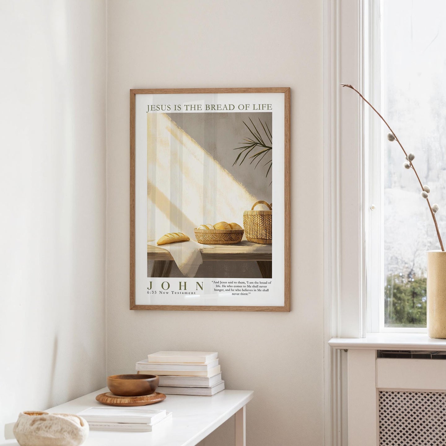 A framed poster with the text "JESUS IS THE BREAD OF LIFE" hangs on a white wall above a white table with a stack of books and a small vase.