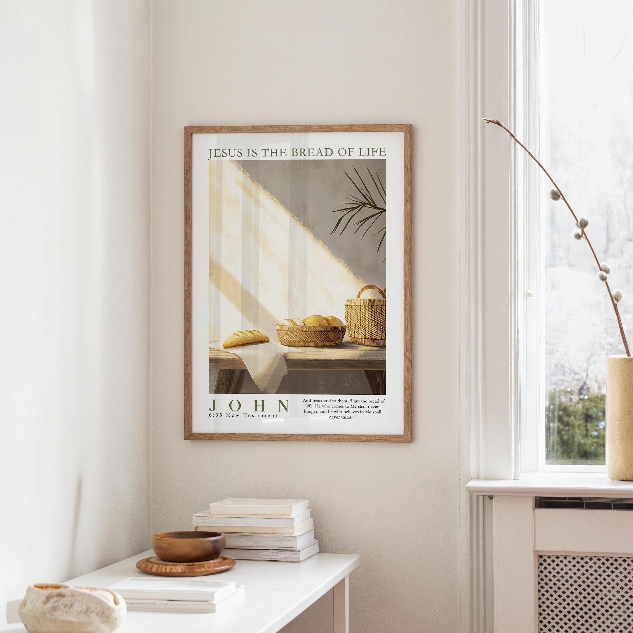 A framed poster with the text "JESUS IS THE BREAD OF LIFE" hangs on a white wall above a white table with a stack of books and a small vase.