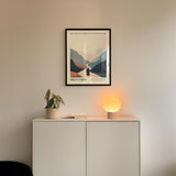 a minimalist interior design with a framed poster of a mountain landscape above a white cabinet with a plant and a lamp on top.