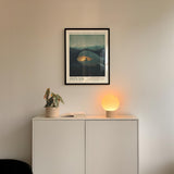 a minimalist interior design with a framed poster of a landscape, a potted plant, and a small lamp on a white cabinet.