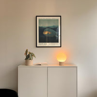 a minimalist interior design with a framed poster of a landscape, a potted plant, and a small lamp on a white cabinet.