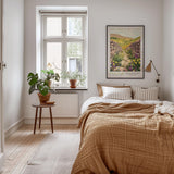 A cozy bedroom with a large bed, a nightstand, and a framed poster of a countryside landscape. The room has a warm, inviting atmosphere with natural elements like plants and a wooden floor.