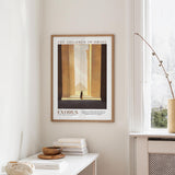 a framed poster of a building with a person standing in the doorway, hanging on a wall in a room with a window and a table with books and a bowl on it.
