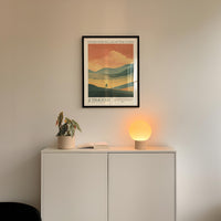 a minimalist interior design with a framed poster of a landscape, a potted plant, and a small lamp on a white cabinet.