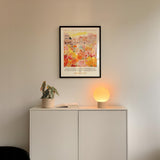 a white cabinet with a potted plant and a lamp on top, and a framed poster of a cityscape hanging on the wall above it.