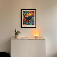 a white cabinet with a plant and a lamp on top, and a framed poster of a colorful whale hanging on the wall above the cabinet.