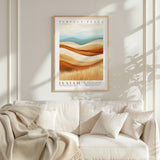 a framed poster of a landscape with a desert scene, hanging on a wall above a white couch with a blanket draped over it.