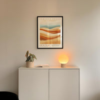 a minimalist interior design with a framed poster of a desert landscape above a white cabinet with a plant and a lamp on top.