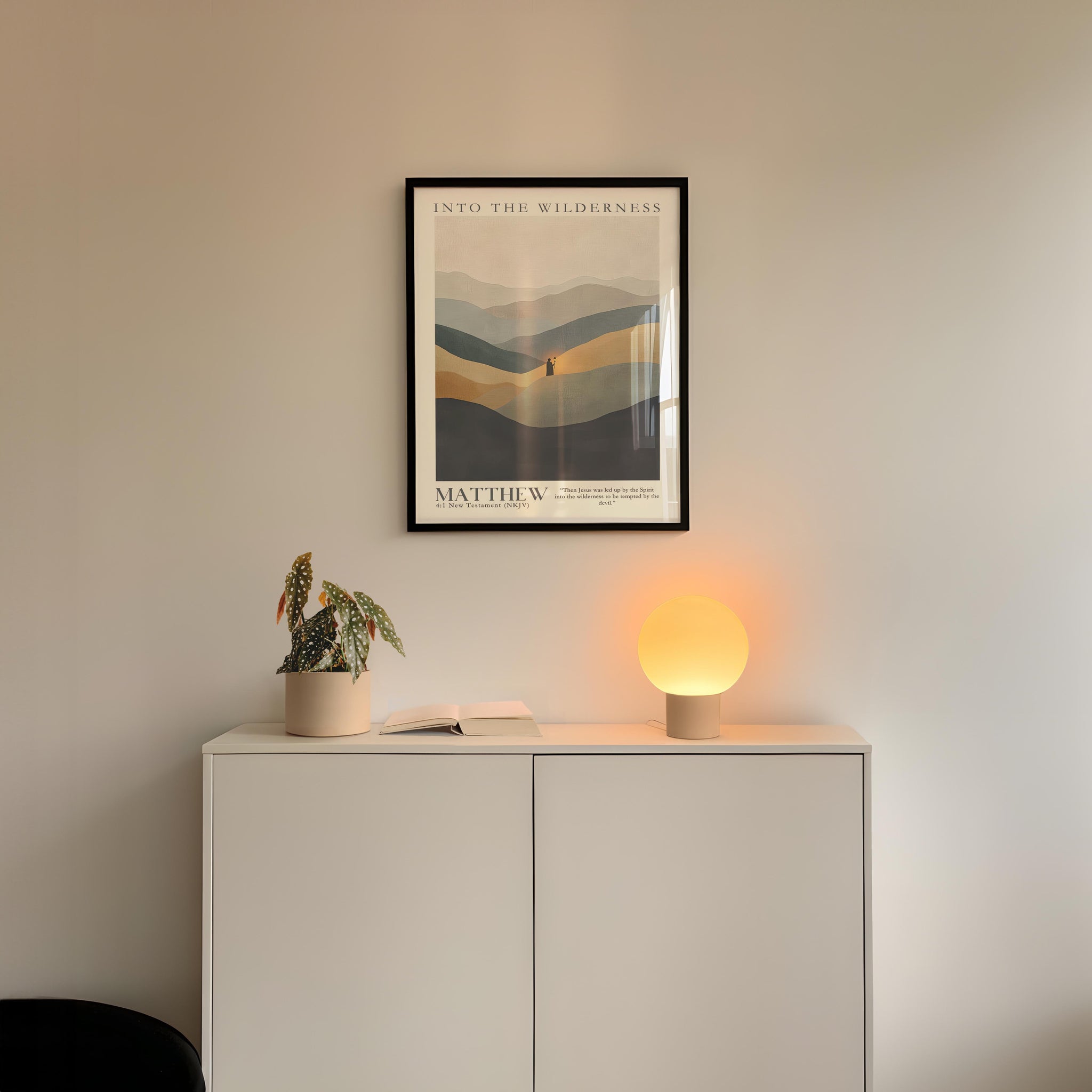 a minimalist interior design with a framed poster of a mountain landscape hanging on a white wall above a white cabinet. The cabinet has a plant and a lamp on top of it, creating a cozy and inviting atmosphere.