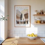 a framed poster of a mountain landscape with a group of people gathered around it, hanging on a wall above a wooden counter with a bowl of lemons.