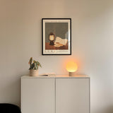 a white cabinet with a plant and a lamp on top, and a framed poster of a lantern and a book on the wall above it.
