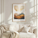 a framed poster of a landscape with mountains and clouds hanging on a wall above a white couch with a blanket draped over it.