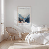 a serene bedroom scene with a bed, a woven chair, and a framed poster of mountains on the wall.
