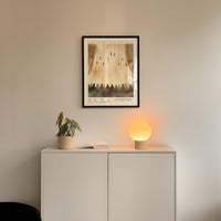a minimalist interior design with a framed poster of a group of people hanging on a white wall above a white cabinet. The cabinet has a plant and a lamp on it, creating a cozy and inviting atmosphere.