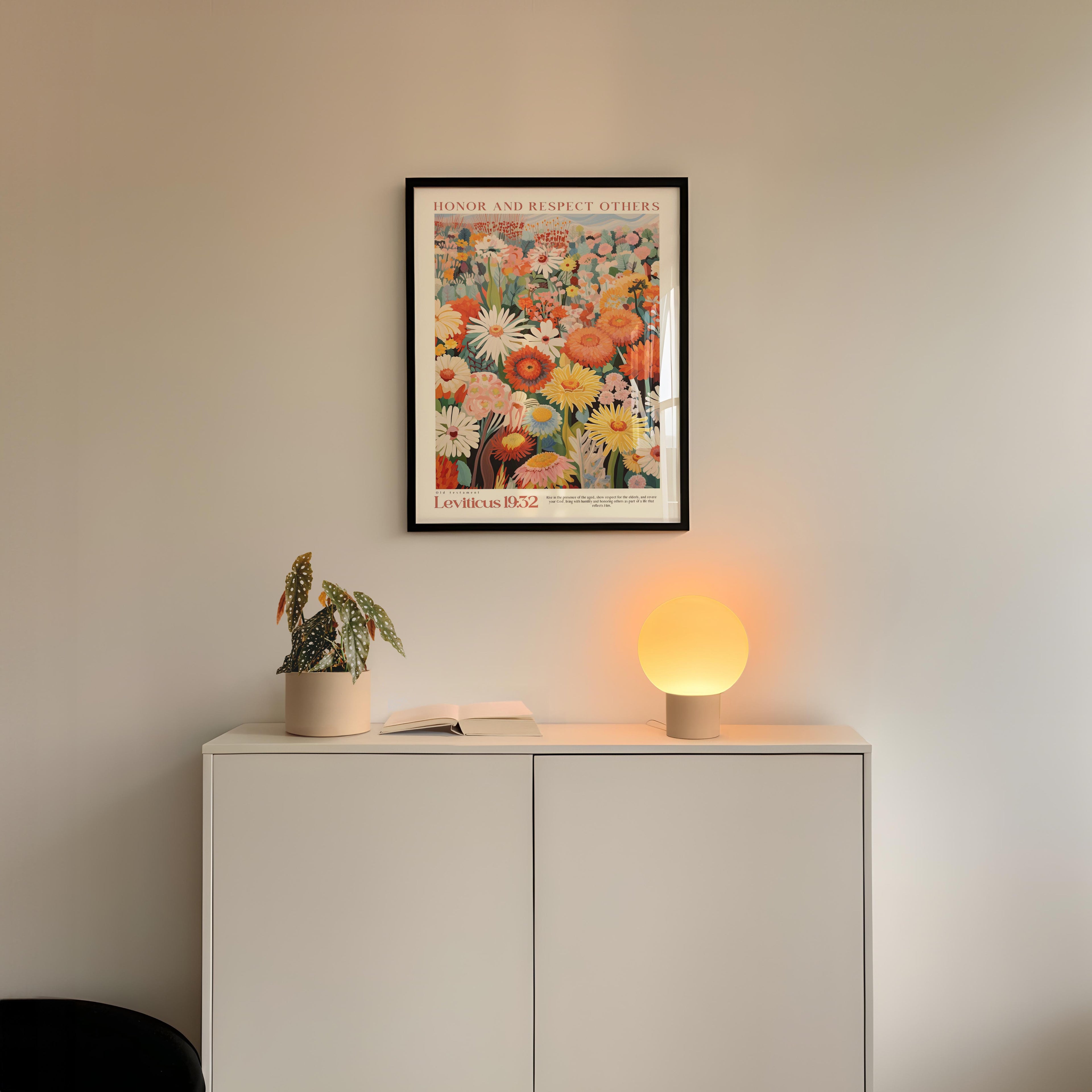a white cabinet with a potted plant and a lamp on top, set against a white wall. Above the cabinet, there is a framed poster of a colorful floral design.