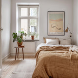 A cozy bedroom with a large bed, a nightstand, and a framed poster on the wall. The room has a warm, inviting atmosphere with natural light coming through a window.