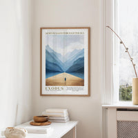 a framed poster of a mountain landscape with a lone figure walking through it, hanging on a wall in a room with a white table and a vase.