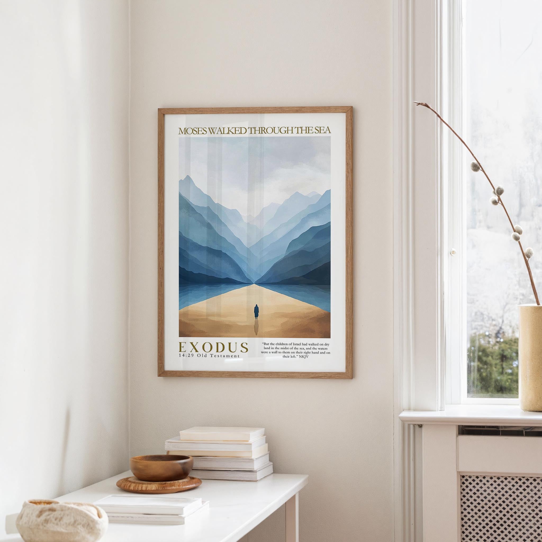 a framed poster of a mountain landscape with a lone figure walking through it, hanging on a wall in a room with a white table and a vase.