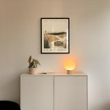 a minimalist interior design with a framed poster of a desert landscape hanging on a white wall above a white cabinet. The cabinet has a plant and a lamp on it, creating a cozy and inviting atmosphere.