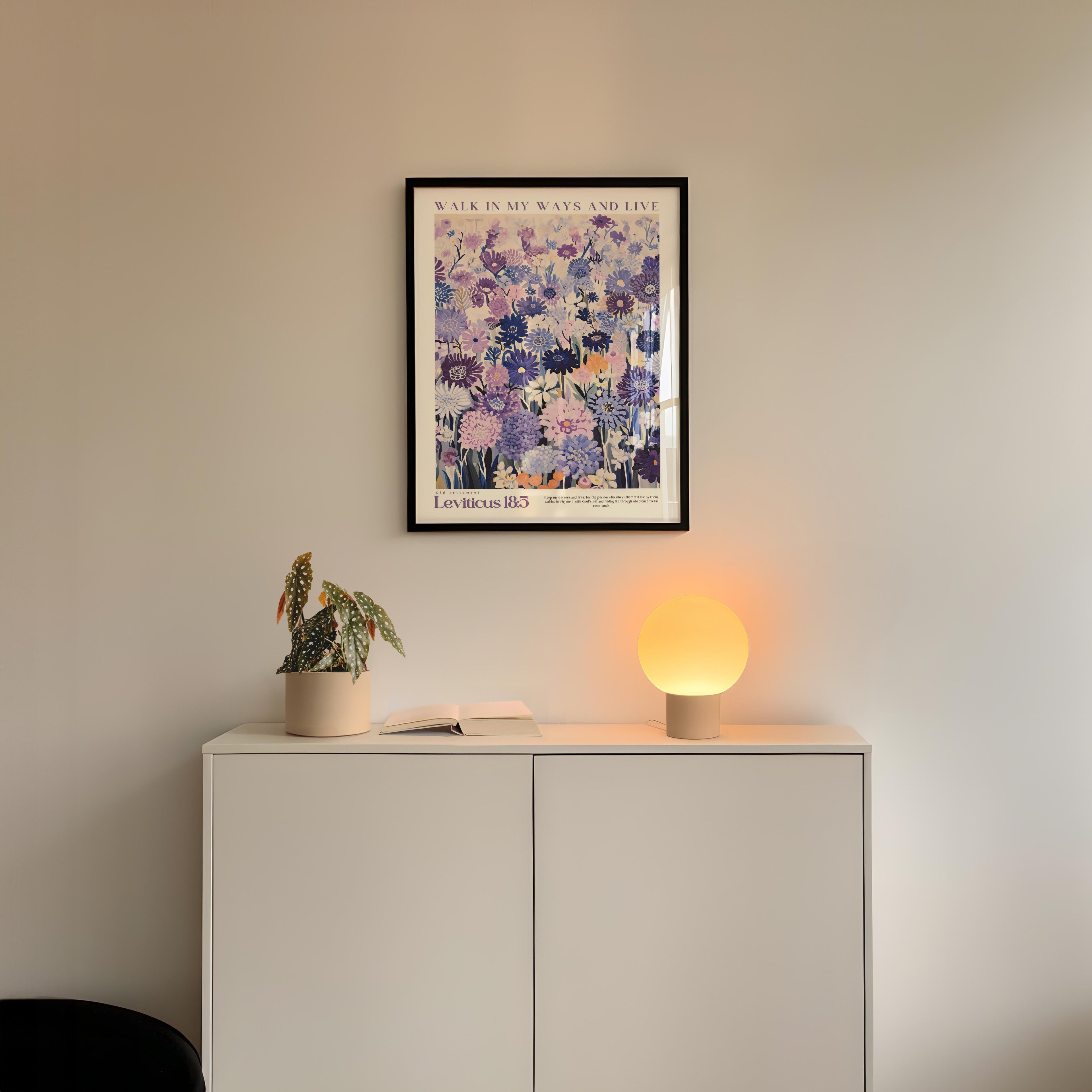 a white cabinet with a potted plant and a lamp on top, set against a white wall. Above the cabinet, there is a framed poster with a floral design.