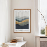 a framed poster of a mountain landscape hanging on a wall, with a stack of books and a vase on a table below it.