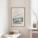 a framed poster of a floral design hanging on a wall, with a window and a small table with books and a bowl below it.