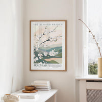 a framed poster of a floral design hanging on a wall, with a window and a small table with books and a bowl below it.