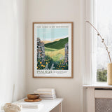 a framed poster of a pastoral landscape with a stone wall and flowers, hanging on a wall next to a window.