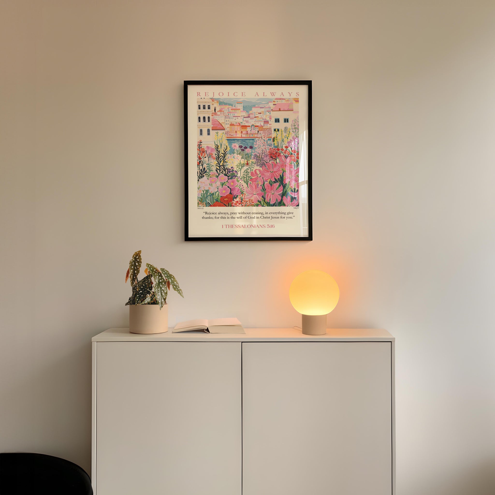 a white cabinet with a potted plant and a lamp on top, set against a white wall. Above the cabinet, there is a framed poster with a colorful illustration of a building and flowers.