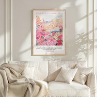 A framed poster of a cityscape with pink flowers hangs on a white wall above a beige couch with a blanket draped over it.