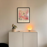 a white cabinet with a potted plant and a lamp on top, set against a white wall. Above the cabinet, there is a framed poster with a floral design.