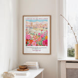 a framed poster of a colorful floral scene hanging on a wall, with a stack of books and a vase on a table below it.