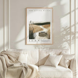 a framed poster of a serene landscape with a well in the center, hanging on a wall above a white couch with a blanket draped over it.