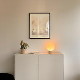 a white cabinet with a plant and a lamp on top, and a framed poster of a landscape hanging on the wall above it.