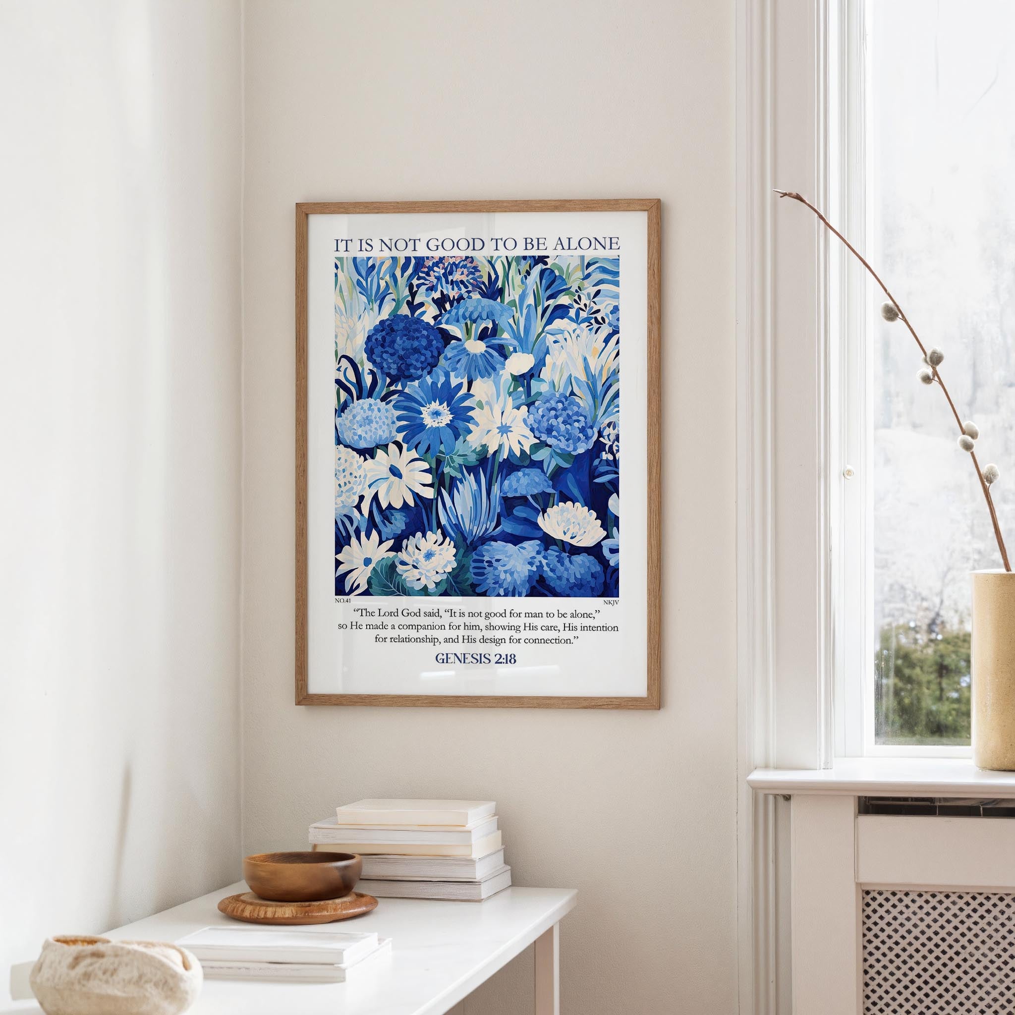 a framed poster of a blue floral design hanging on a white wall, with a stack of books and a vase on a white table below it.