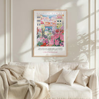 a framed poster of a colorful garden scene hanging on a wall above a white couch with a blanket draped over it.