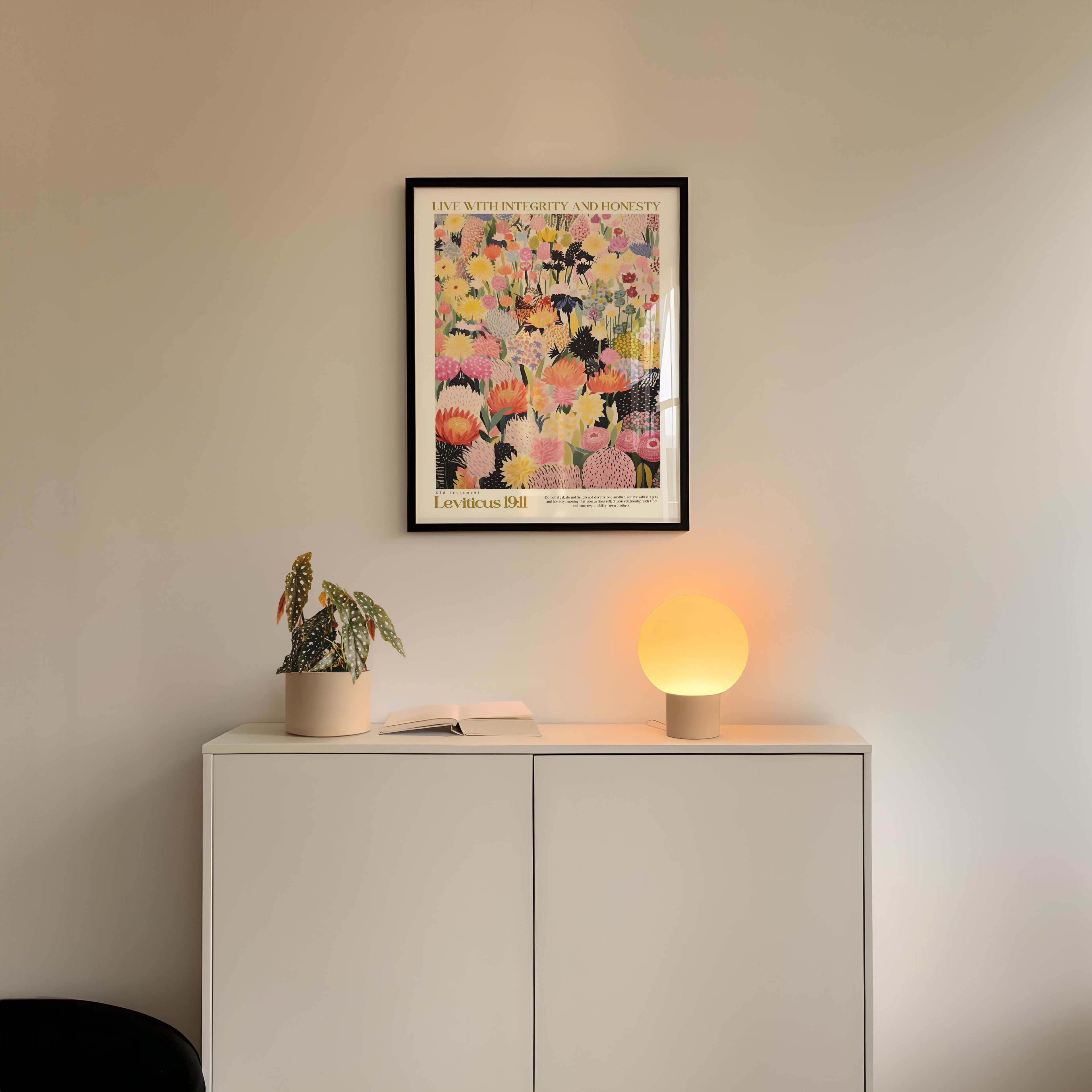 a white cabinet with a potted plant and a lamp on top, set against a white wall. Above the cabinet, there is a framed poster with a colorful floral design.