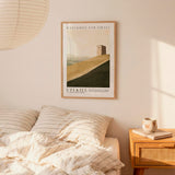 a bedroom setting with a framed poster of a building on the wall, a bed with striped bedding, and a wooden nightstand with a coffee mug and a book.