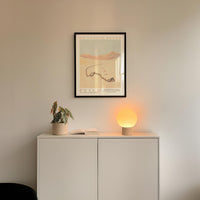 a white cabinet with a potted plant and a lamp on top, and a framed poster of a desert landscape hanging on the wall above the cabinet.