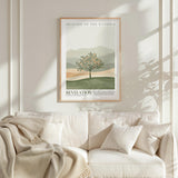 a framed poster of a tree with a landscape in the background, hanging on a wall above a white couch with pillows.