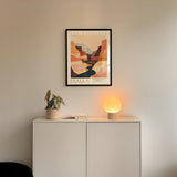 a minimalist interior design with a framed poster of a desert landscape hanging on a white wall above a white cabinet. The cabinet has a plant and a lamp on it, creating a cozy and inviting atmosphere.