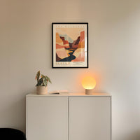 a minimalist interior design with a framed poster of a desert landscape hanging on a white wall above a white cabinet. The cabinet has a plant and a lamp on it, creating a cozy and inviting atmosphere.